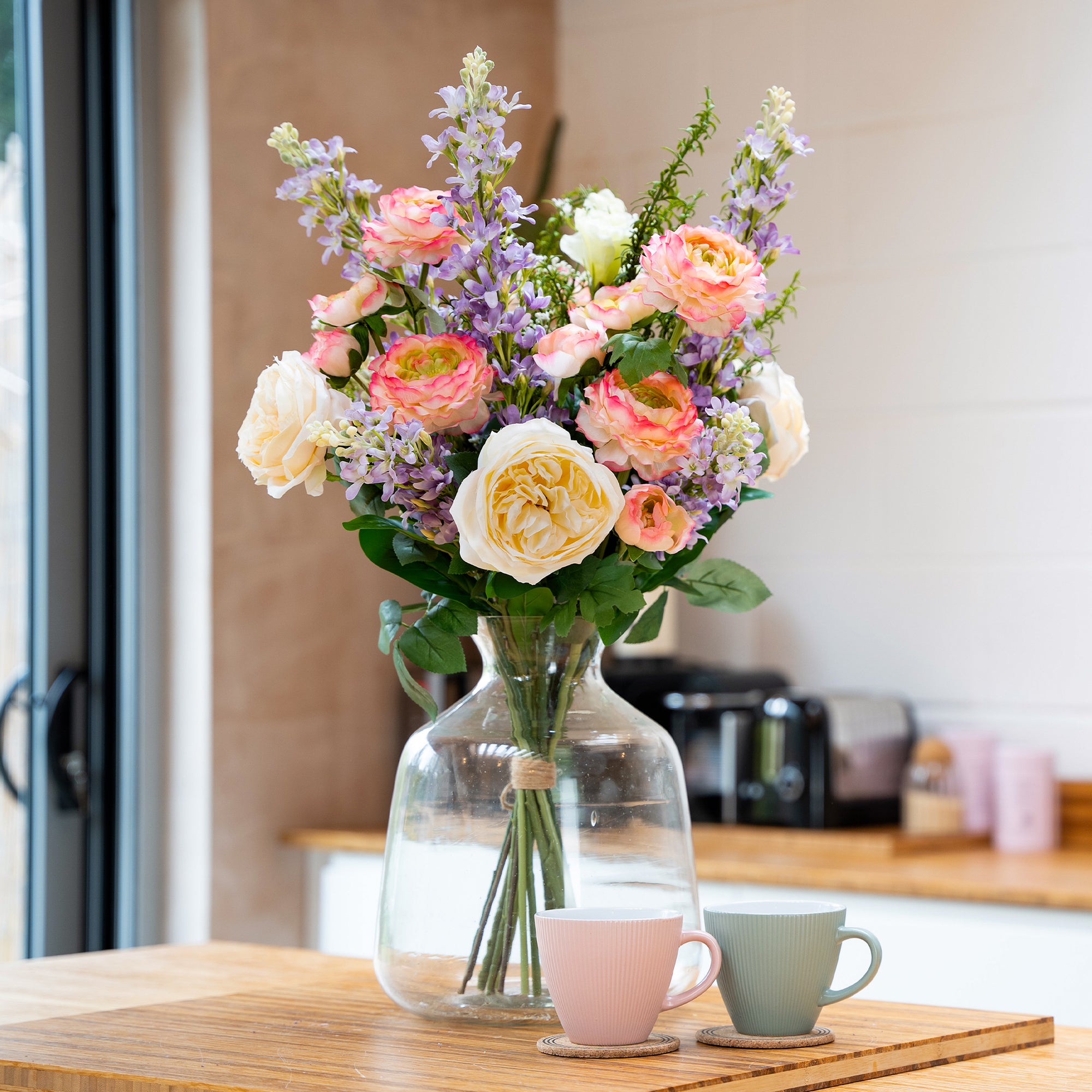 Bouquet of flowers in a clear vase on a wooden counter with two cups in a kitchen setting.