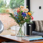 Colorful bouquet of flowers in a vase on a wooden table with a couch and window in the background