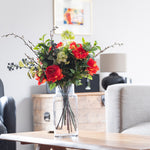 Bouquet of red and green flowers in a clear vase on a wooden table in a living room.