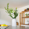 White vase with greenery and white flowers on a table in a room with wooden shelves.