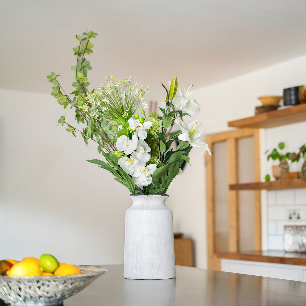 White vase with greenery and white flowers on a table in a room with wooden shelves.