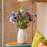 Vase with flowers on a table next to a lamp and colorful cushions on a couch.