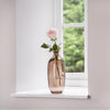 Vase with a single pink rose on a windowsill with a blurred green outdoor background