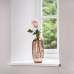 Vase with a single pink rose on a windowsill with a blurred green outdoor background