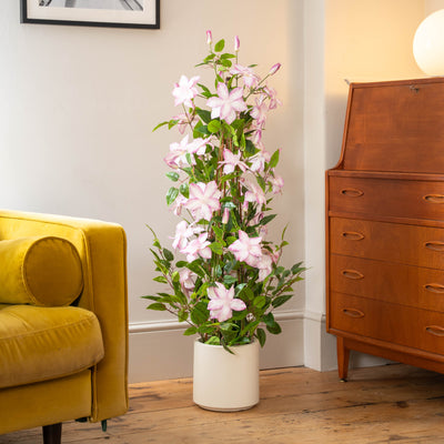 A tall potted artificial flowering plant with light pink flowers and green leaves, displayed in a white cylindrical pot on a wooden floor next to a yellow armchair and a mid-century modern wooden cabinet.