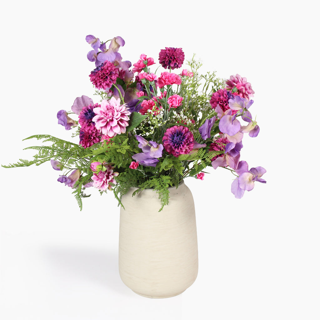 A vibrant artificial flower bouquet featuring pink, purple, and magenta blossoms with green foliage, arranged in a textured cream-colored ceramic vase against a plain white background.