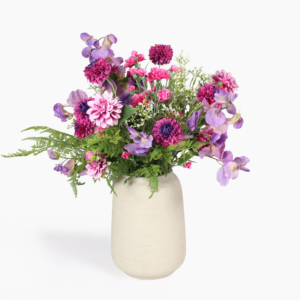 A vibrant artificial flower bouquet featuring pink, purple, and magenta blossoms with green foliage, arranged in a textured cream-colored ceramic vase against a plain white background.
