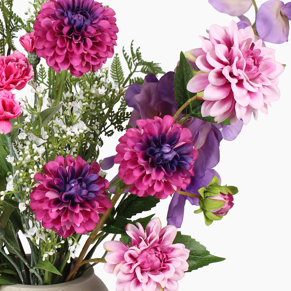 A close-up of a mixed floral arrangement featuring vibrant pink and purple dahlia flowers with green foliage, arranged in a light-colored vase against a plain white background.