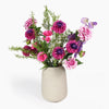 A faux floral arrangement featuring pink, purple, and magenta flowers with green foliage, presented in a textured light beige ceramic vase against a clean white background.