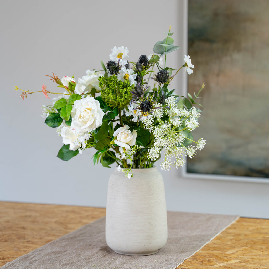 A faux flower arrangement featuring white roses, white daisies, green foliage, and dark thistle-like blooms in a textured light grey ceramic vase, set on a wooden table with a neutral background.