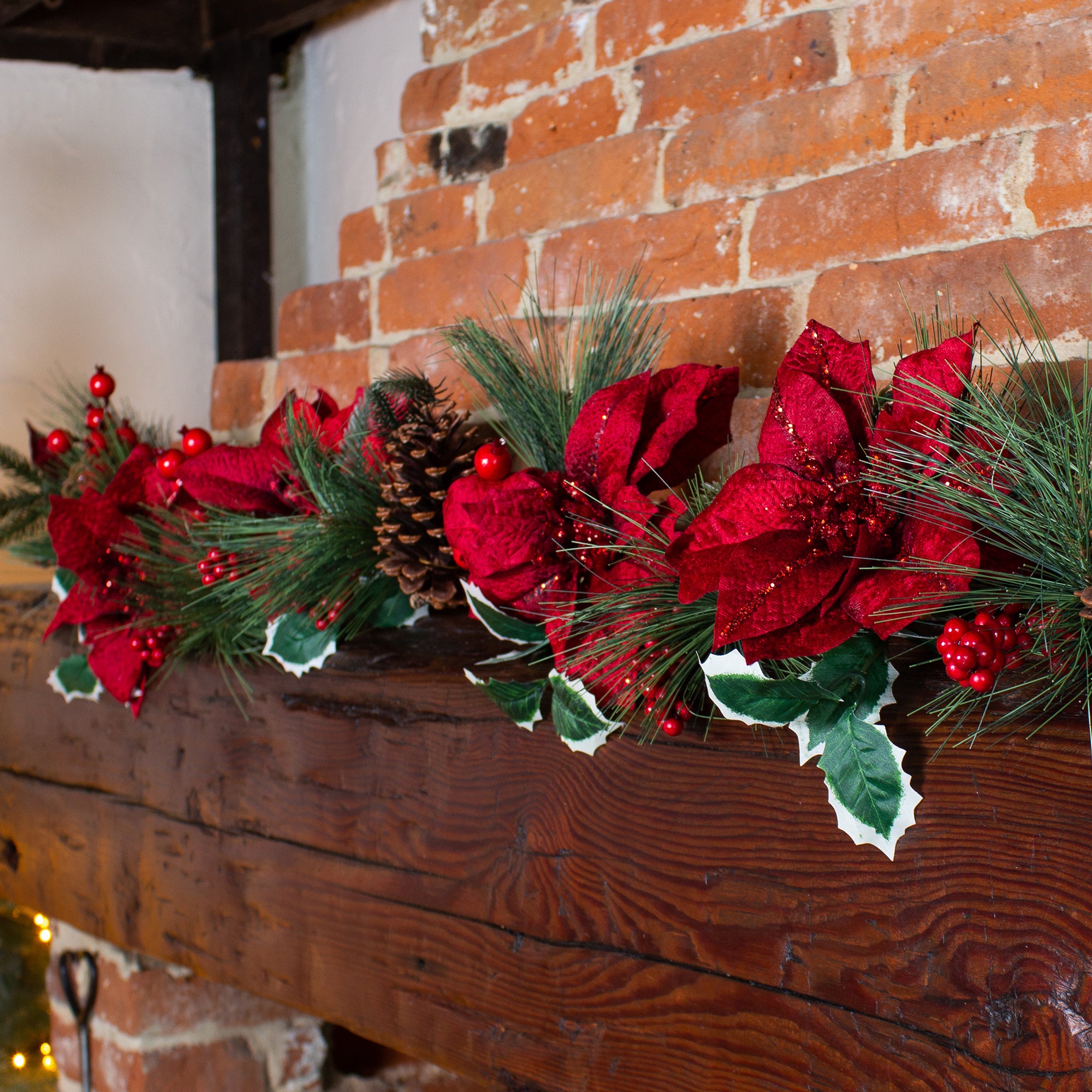 Decorative Christmas garland with red flowers and green leaves on a wooden mantel against a brick wall.
