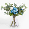 Bouquet of blue hydrangeas and greenery in a clear glass vase on a white background