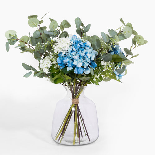 Bouquet of blue hydrangeas and greenery in a clear glass vase on a white background