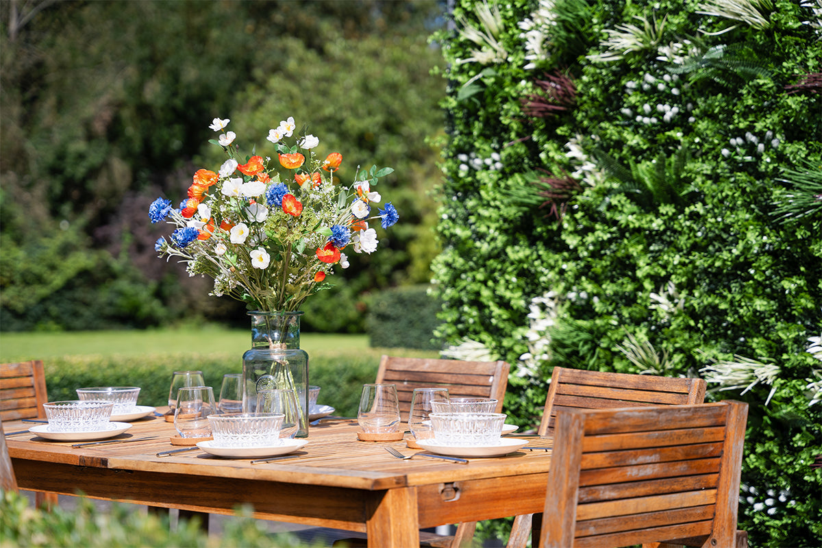 Outdoor dining setup with wooden table and chairs, flowers in a vase, and greenery in the background.