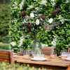 Bouquet of purple and white flowers in a clear vase on a wooden table outdoors.