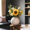 Floral arrangement in a white vase on a wooden stool in a living room setting.