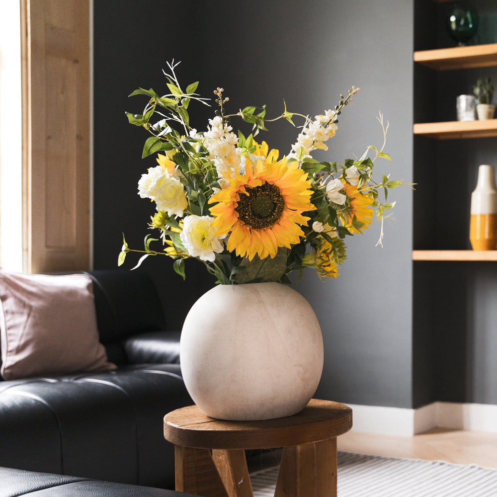 Floral arrangement in a white vase on a wooden stool in a living room setting.