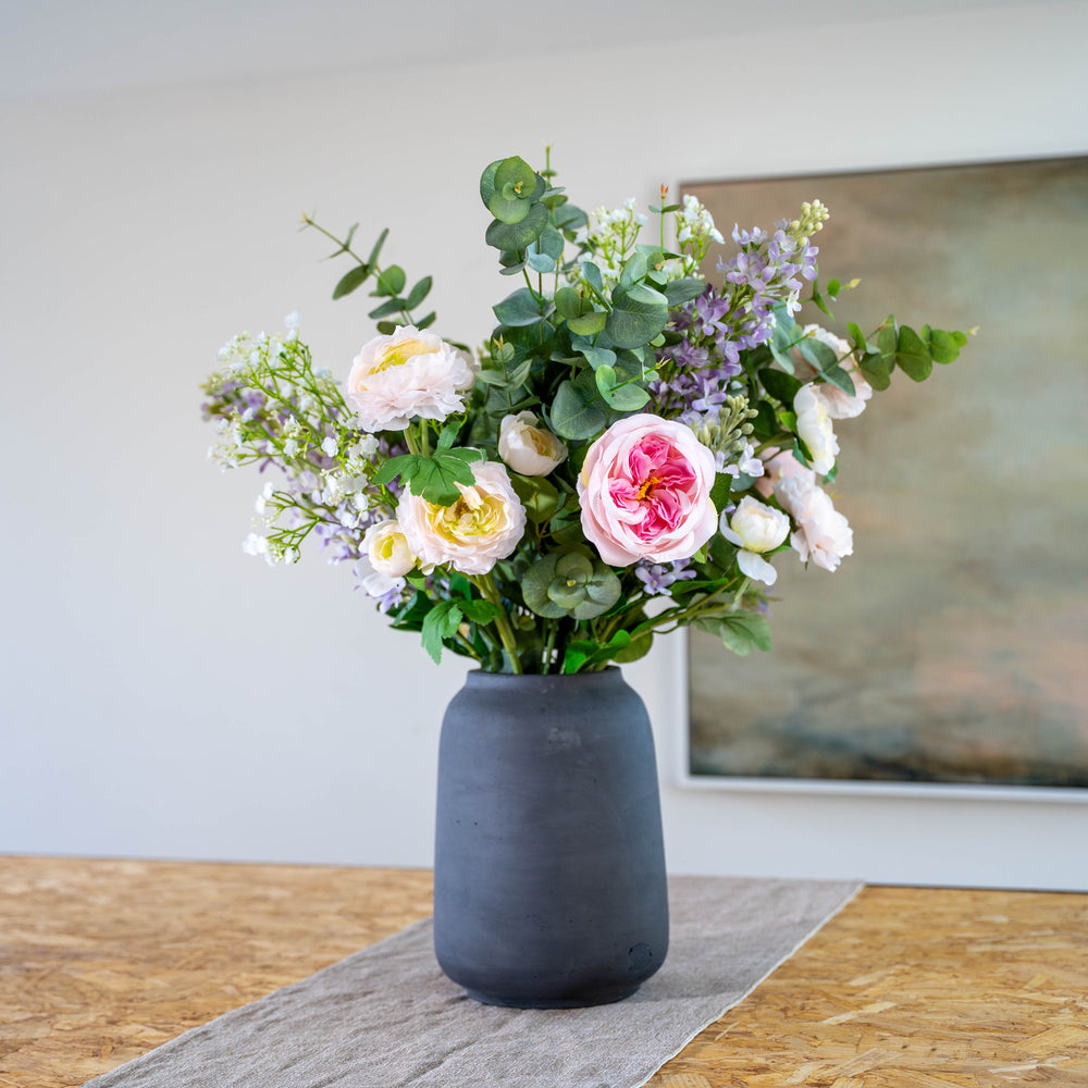 A faux flower arrangement featuring soft pinks, purples and greens in a matte dark gray ceramic vase, placed on a textured runner on a wooden table with a neutral wall and framed art in the background.