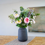 A faux flower arrangement featuring soft pinks, purples and greens in a matte dark gray ceramic vase, placed on a textured runner on a wooden table with a neutral wall and framed art in the background.