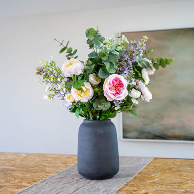 A faux flower arrangement featuring soft pinks, purples and greens in a matte dark gray ceramic vase, placed on a textured runner on a wooden table with a neutral wall and framed art in the background.