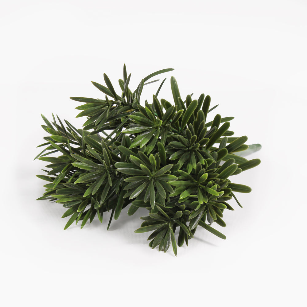 A small artificial green plant with dense, needle-like leaves, photographed on a clean white background.