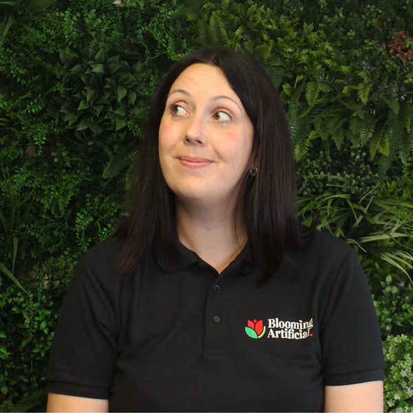 Person wearing a black polo shirt with a 'Blooming Artificial' logo against a green leafy background