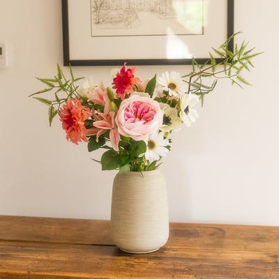 A ceramic vase filled with a colorful white and pink artificial flower arrangement, placed on a wooden table against a white wall with framed art.