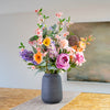 An artificial floral arrangement featuring soft pink, orange, purple and blue flowers and seed heads with sage green foliage, presented in a matte dark gray ceramic vase on a table runner.