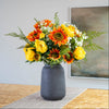 A large and vibrant autumn faux floral arrangement featuring yellow and orange flowers with green foliage, presented in a matte dark gray ceramic vase on a wooden table.