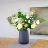 A large faux flower arrangement featuring white, cream, and pale yellow flowers with green woodland foliage, displayed in a textured dark grey ceramic vase on a table runner.