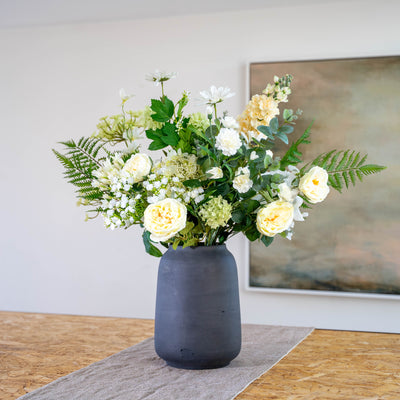 A large faux flower arrangement featuring white, cream, and pale yellow flowers with green woodland foliage, displayed in a textured dark grey ceramic vase on a table runner.