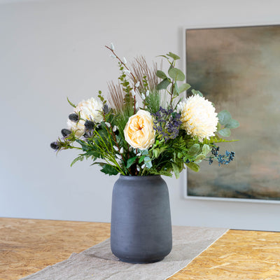 An artificial white flower and green foliage arrangement in a matte dark gray ceramic vase, placed on a beige runner over a wooden table with a neutral wall and framed artwork in the background.